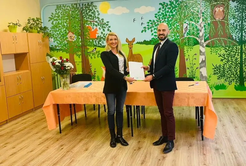 Zdjęcie o orientacji poziomej przedstawia dwie osoby uśmiechnięte do obiektywu, stojące w sali przedszkolnej. Po lewej stronie znajduje się blondynka w czarnej marynarce i jeansach, a po prawej mężczyzna w garniturze, którzy wspólnie trzymają białą kartkę papieru. Przed nimi stoi stół nakryty pomarańczowym obrusem, na którym widać bukiet kwiatów oraz inne przedmioty. Tło stanowi kolorowy mural przedstawiający leśne zwierzęta, takie jak sarna, sowa i wiewiórka, oraz liczne drzewa. Po lewej stronie kadru widoczne są drewniane szafki, a podłoga wykonana jest z paneli imitujących jasne drewno.