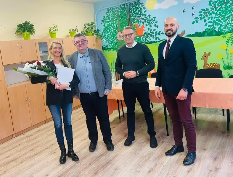 Zdjęcie przedstawia grupę czterech osób pozujących w jasnym pomieszczeniu, prawdopodobnie w sali przedszkolnej, w formacie pionowym o proporcjach zbliżonych do 4:5. Po lewej stronie stoi kobieta w czarnej marynarce, trzymająca bukiet kwiatów oraz dokument, a obok niej stoi trzech mężczyzn w strojach od eleganckich po biznesowe. W tle widać ściany ozdobione kolorowym malowidłem przedstawiającym przyrodę, las i zwierzęta, co sugeruje placówkę oświatową. Po lewej stronie znajdują się wysokie, drewniane szafki, a na pierwszym planie stoi stół przykryty brzoskwiniowym obrusem. Całość kompozycji jest symetryczna, z postaciami ustawionymi w rzędzie, co pozwala na czytelne przedstawienie wszystkich osób w przestrzeni oświetlonej równomiernym światłem dziennym.