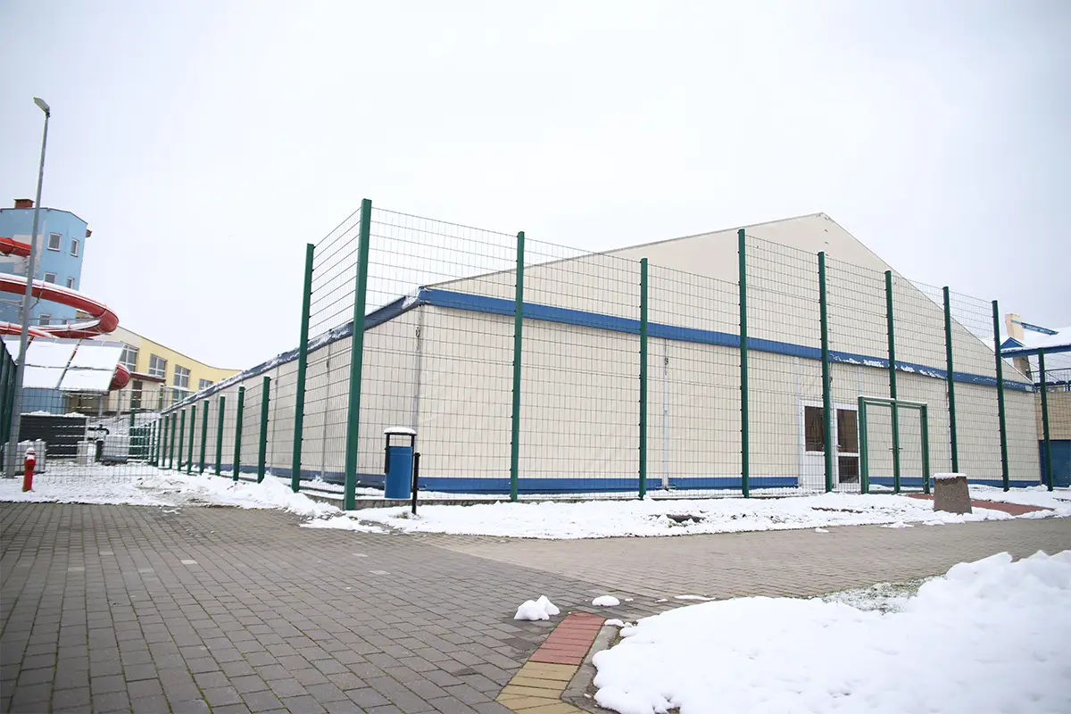 Zdjęcie przedstawia dużą halę namiotową o jasnych ścianach i dwuspadowym dachu, która jest otoczona wysokim metalowym ogrodzeniem z zielonymi słupkami. Obiekt o proporcjach panoramicznych wkomponowany jest w otoczenie wyłożone szarą kostką brukową, na której miejscami zalega warstwa białego śniegu. W tle po lewej stronie widoczna jest część niebieskiego budynku z czerwoną, spiralną zjeżdżalnią, co sugeruje lokalizację w pobliżu parku wodnego lub ośrodka rekreacyjnego. Przy ogrodzeniu ustawiony jest pojedynczy, niebieski kosz na śmieci, a po prawej stronie znajduje się betonowy element małej architektury. Całość kompozycji utrzymana jest w chłodnej, zimowej kolorystyce, gdzie surowość technicznej konstrukcji kontrastuje z zaśnieżonym terenem.