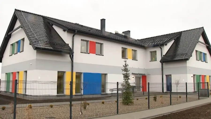 Zdjęcie przedstawia nowoczesny budynek użyteczności publicznej lub przedszkole w układzie poziomym o proporcjach boków 16:9. Biała elewacja obiektu została urozmaicona szarym pasem biegnącym przez środek oraz licznymi kolorowymi wstawkami w odcieniach czerwieni, niebieskiego, żółtego i zielonego przy oknach. Budynek posiada dwuspadowy dach pokryty ciemną dachówką oraz liczne okna o nowoczesnym profilu. Całość otoczona jest metalowym ogrodzeniem panelowym typu siatka, za którym widać zadbany teren z młodymi nasadzeniami drzew i iglaków. Przed budynkiem rozciąga się chodnik z kostki brukowej, a wejście główne wyposażone jest w podjazd dla osób niepełnosprawnych.